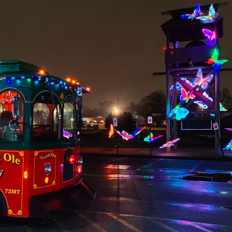 Red and green trolley with colorful lights near glowing butterfly sculptures at night.