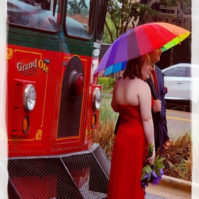 Woman in red dress with rainbow umbrella stands near a red bus.