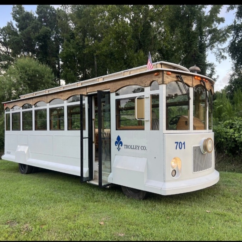White trolley bus with an American flag and logo, parked on grass with trees in the background.