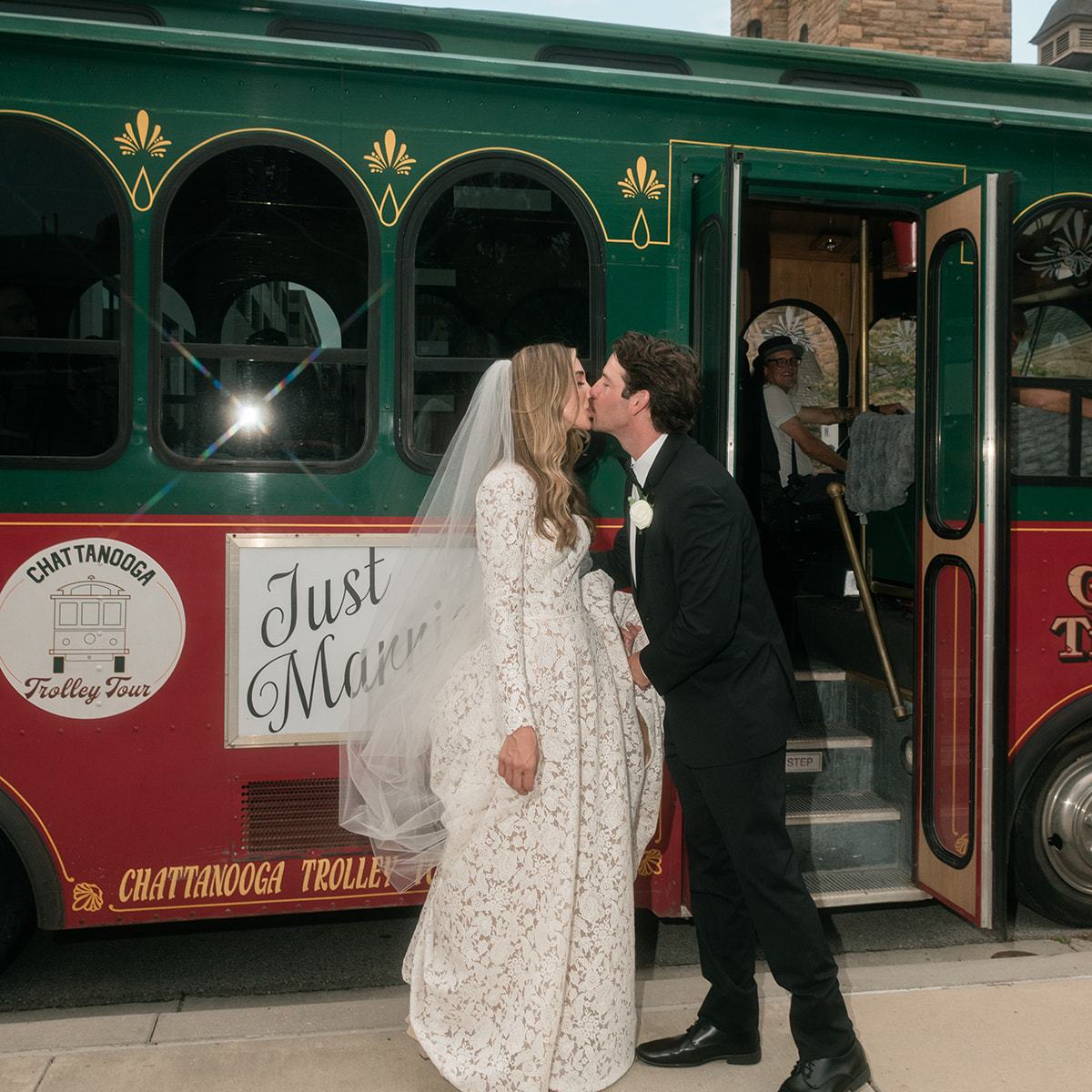 Bride and groom kiss in front of a 'Just Married' trolley, with city buildings in the background.