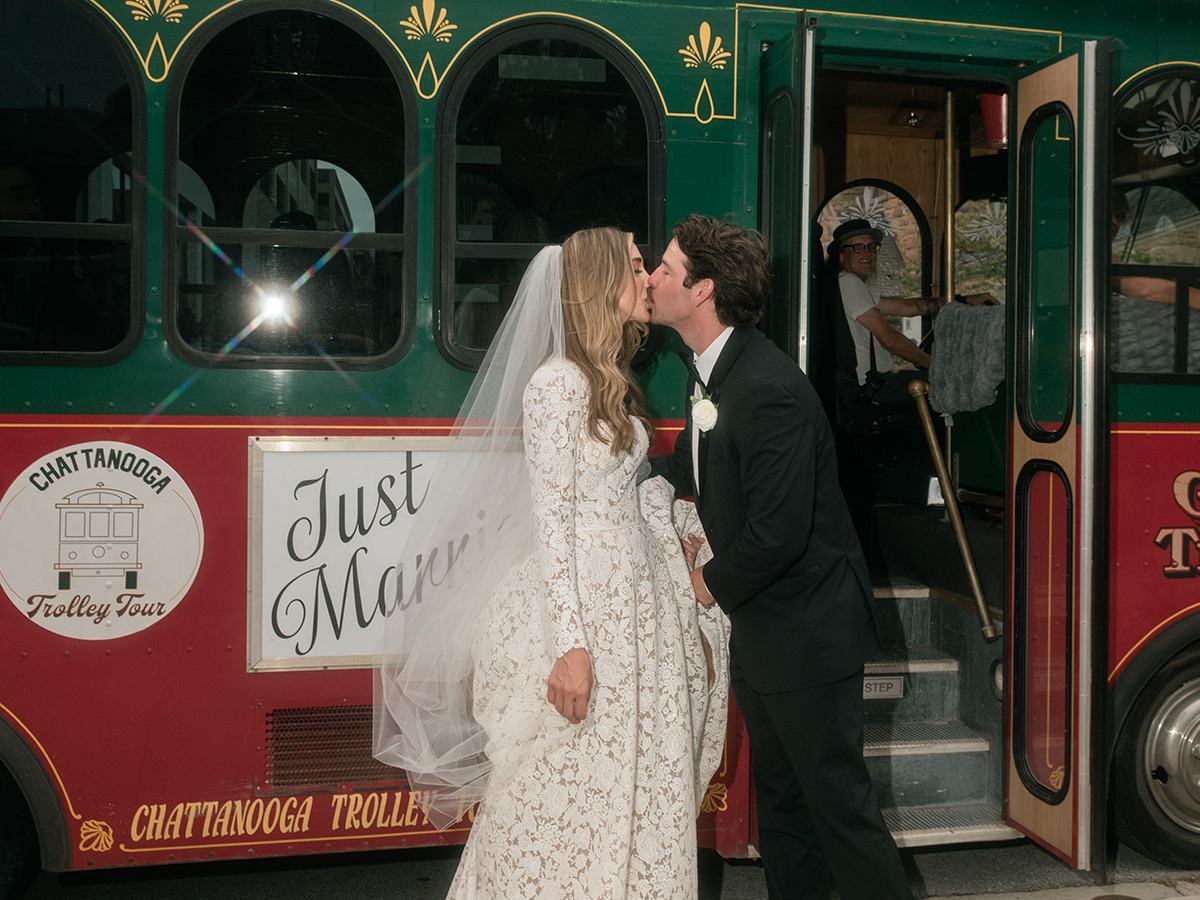 Bride and groom kiss in front of a 'Just Married' trolley, with city buildings in the background.